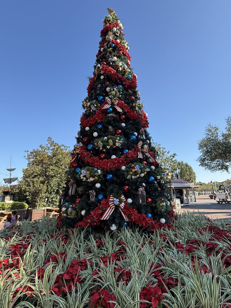 A large, decorated Christmas tree surrounded by red flowers and green foliage, under a clear blue sky.
