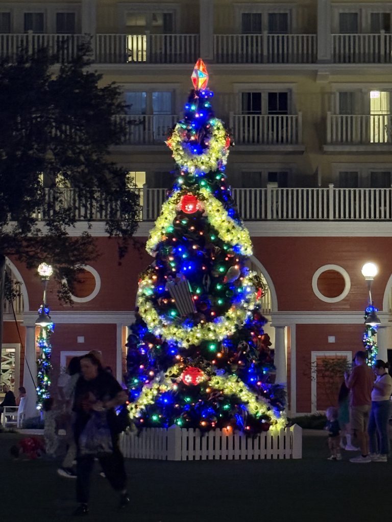 A beautifully decorated Christmas tree illuminated with colorful lights and ornaments, set in a festive outdoor scene at dusk. People stroll by, enjoying the holiday atmosphere.