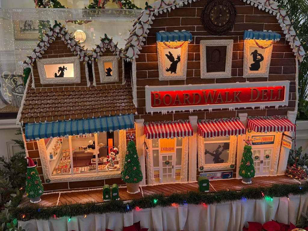A festive gingerbread replica of 'BoardWalk Deli' featuring detailed decorations, colorful awnings, and illuminated windows, surrounded by small Christmas trees and holiday lights.